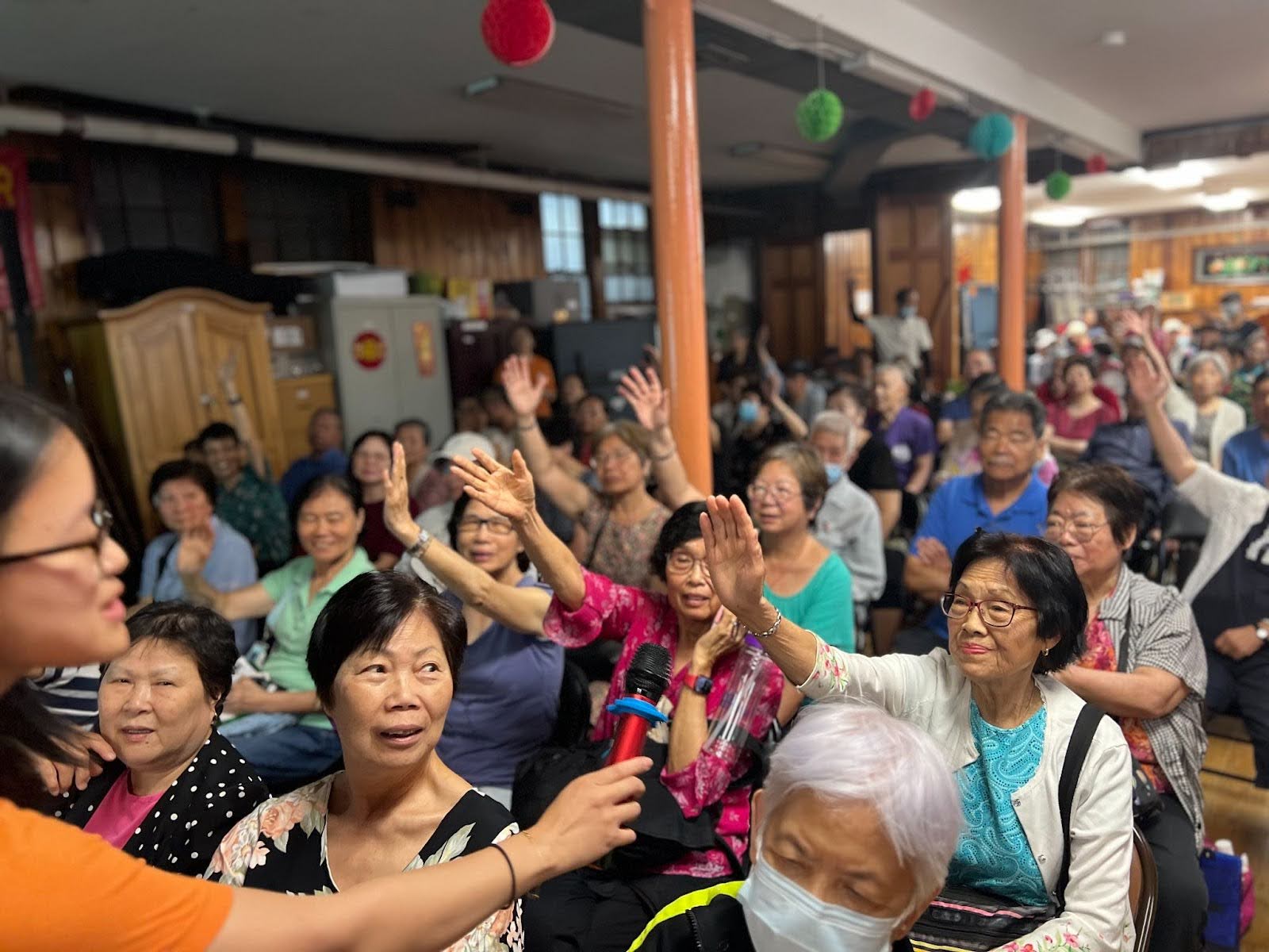 Un groupe de personnes âgées d'origine asiatique assises sur des chaises, parmi lesquelles plusieurs femmes lèvent la main au centre.